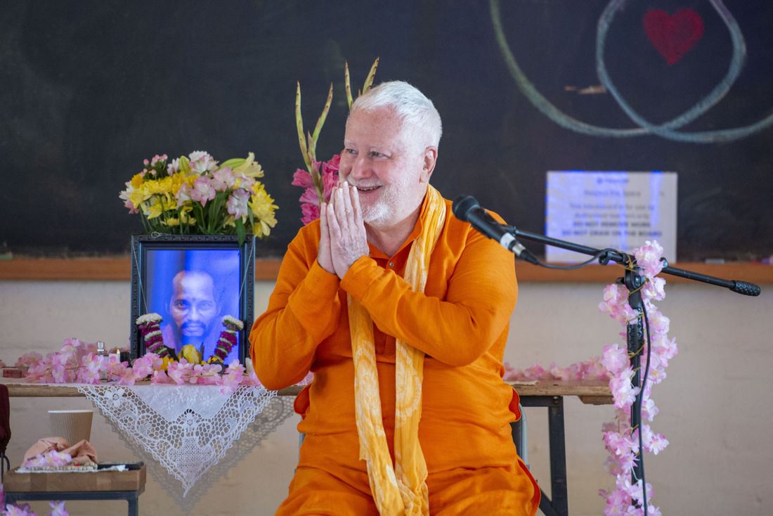 Swami Brahmananda greeting people with his hands together, seated in orange robes beside flowers and a microphone