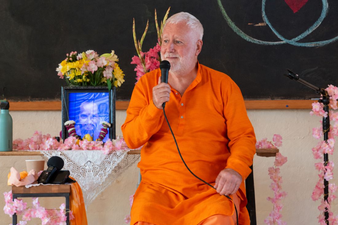 Swami Brahmananda speaking into a microphone, seated beside flowers